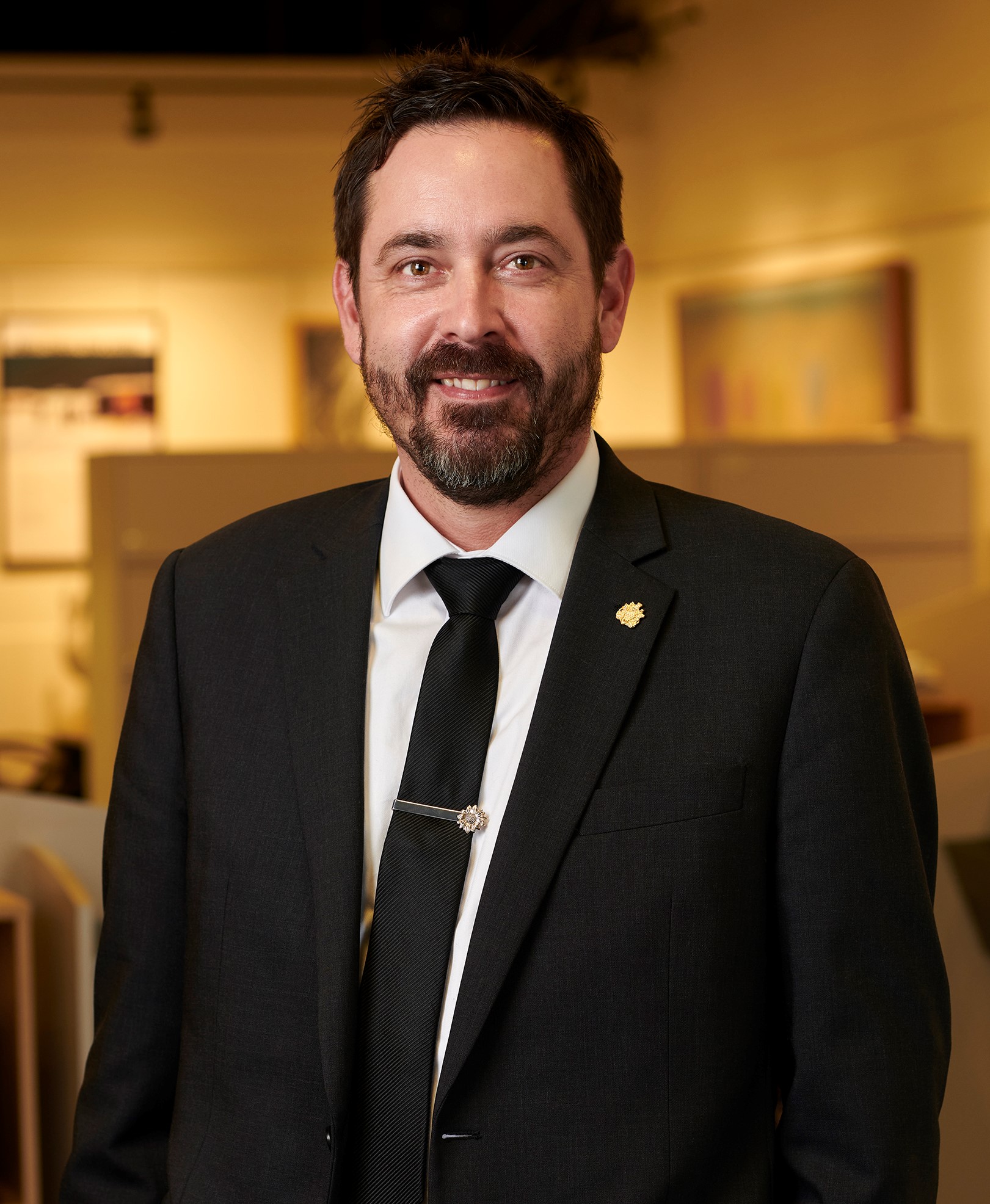 Tall, white male with brown hair and a beard wearing a black suit. He is standing in an office with a blurred background smiling.