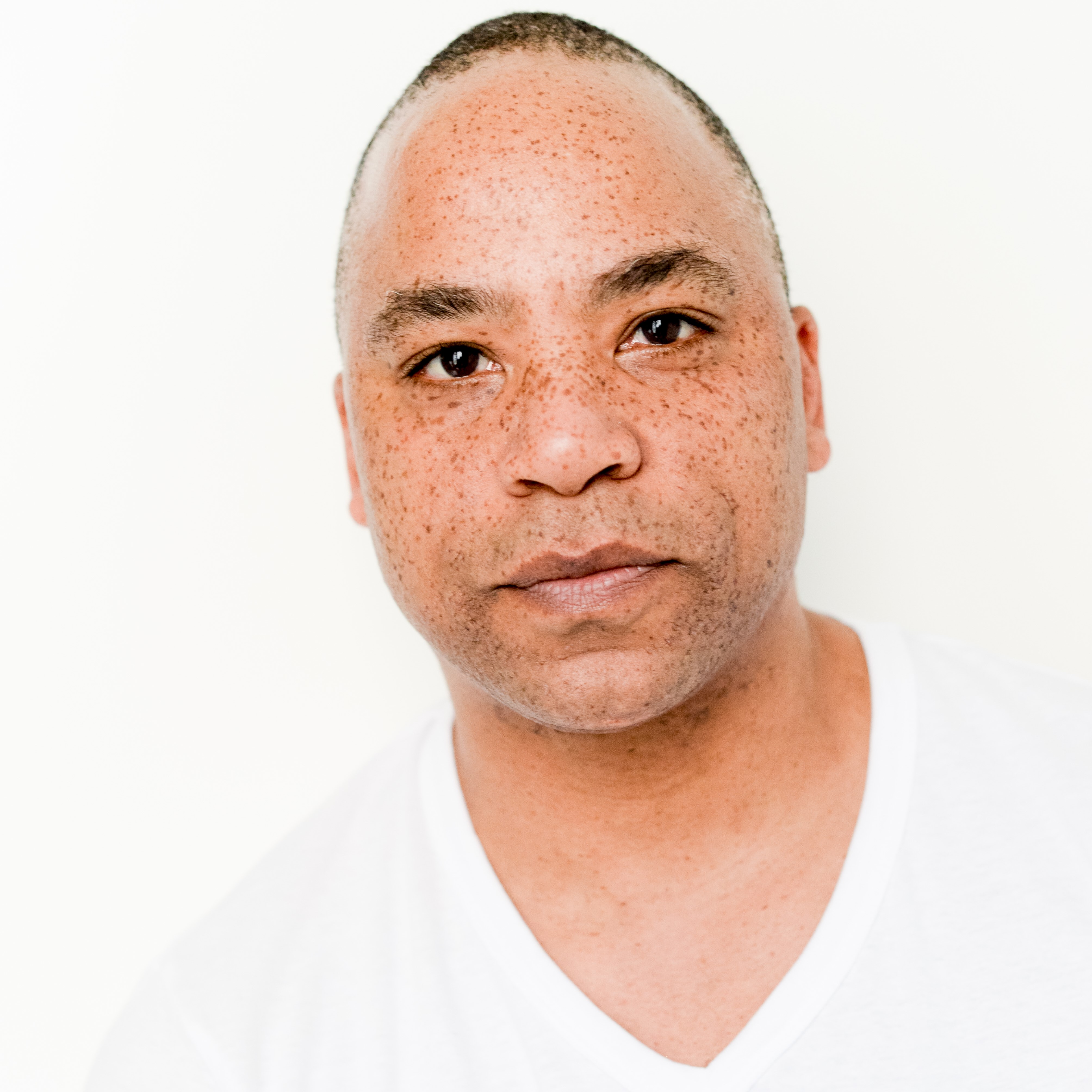 Chancz Perry joins SK Arts- Headshot of black man with freckles wearing a white v neck t-shirt and a white background.