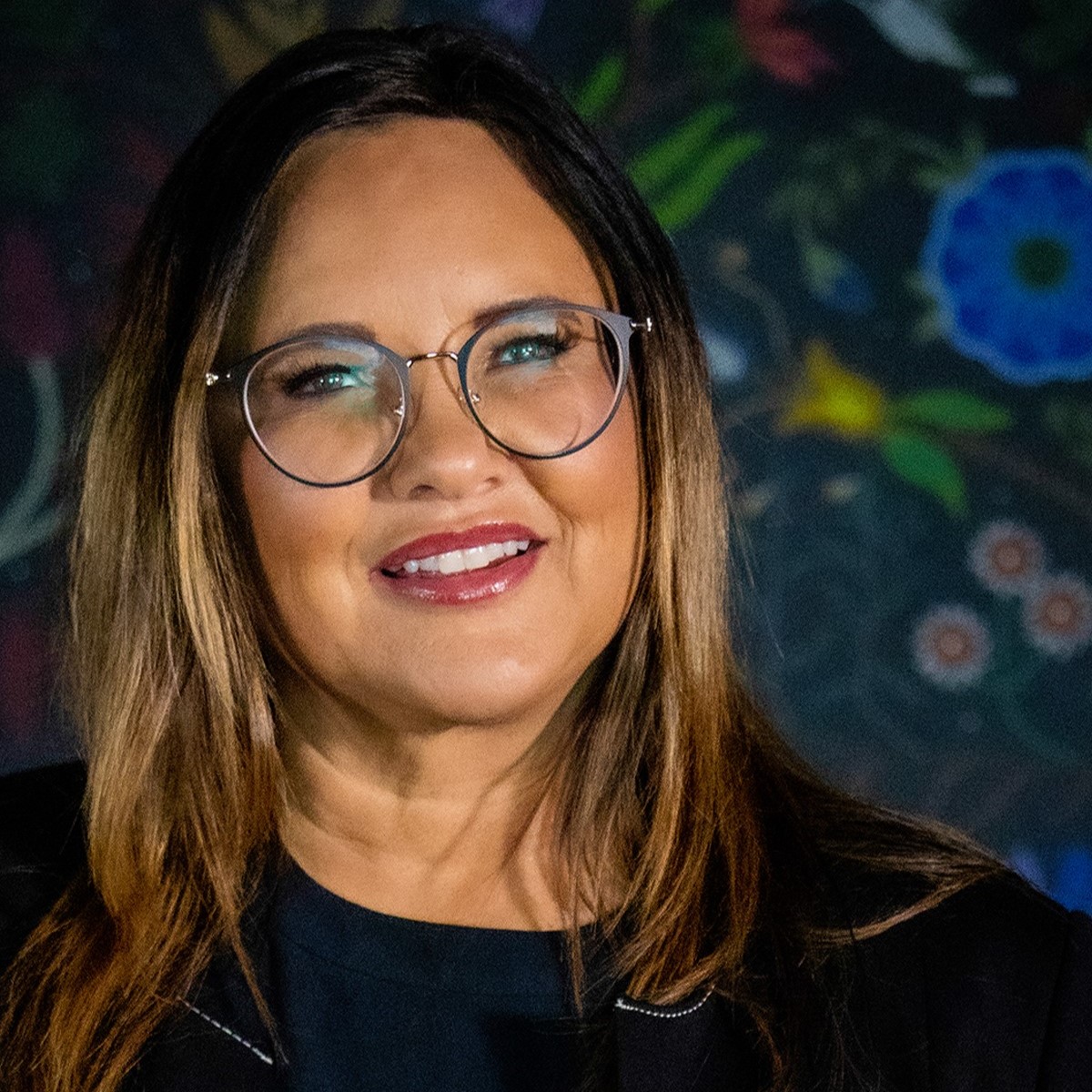 Portrait photo of Indigenous woman with brown and gold Ombre hair wearing round glasses, a black blouse and smiling. She is wearing red lipstick and the background is Indigenous art.  Lisa Bird-Wilson, SK Arts CEO