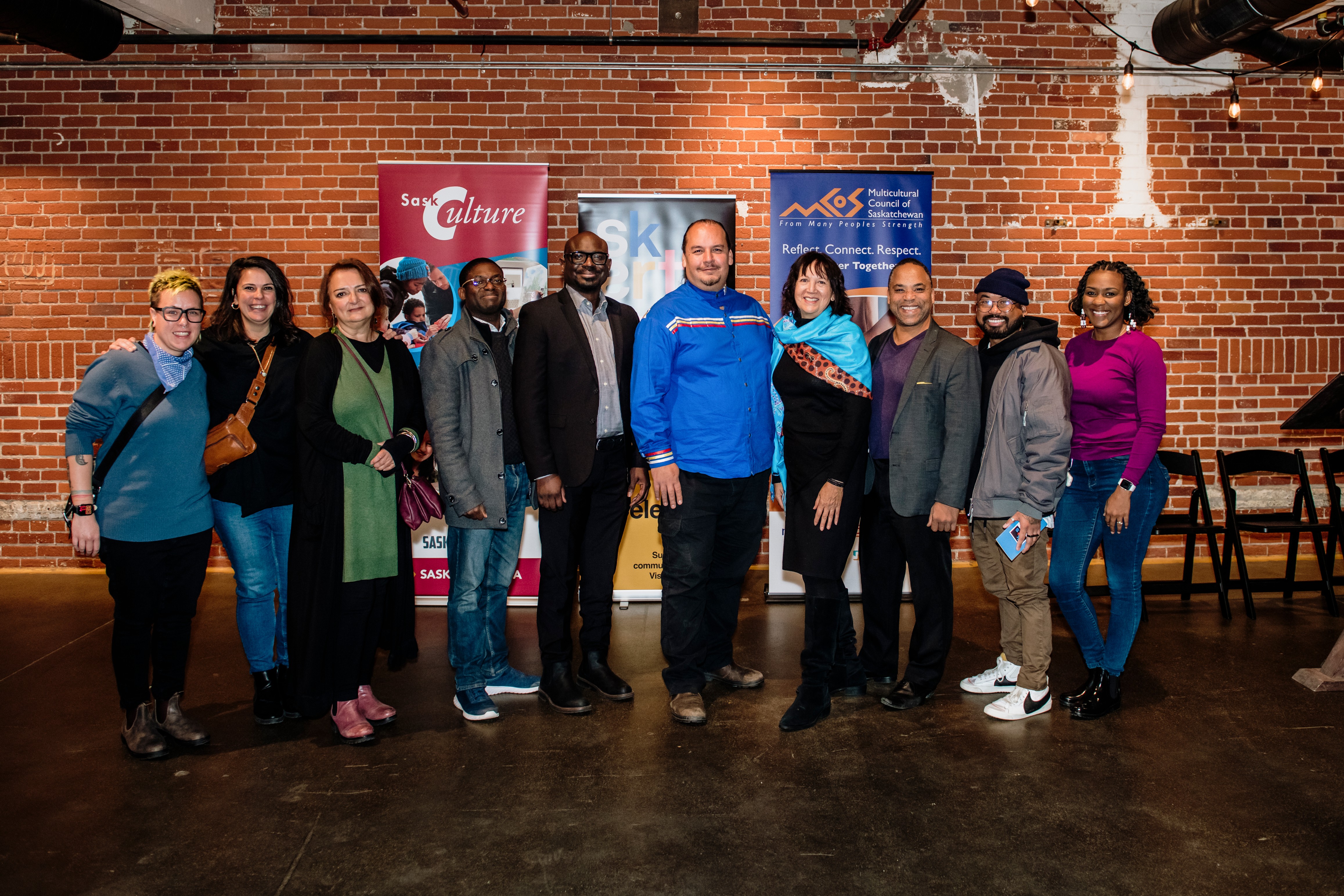 Photo featuring 10 people standing in front of a brick wall and banners for Sask Culture, SK Arts and the Multicultural Society of Saskatchewan. In the photo (L-R): Em Ironstar (Saskatchewan Arts Alliance executive director), Muveddet Al-Katib, Anahit Falihi, Dr. Taiwo Afolabi, Michael Afenfia (SK Arts Board member), Damon Badger Heit (SaskCulture supervisor for partnership and inclusion), Carol Greyeyes (SK Arts Policy and Planning Advisor), Chancz Perry (dancer), Eddy Alvaro (dancer), Shandean Reid (SK Arts communications strategist) at the Building Arts Equity POC Program launch in Regina.