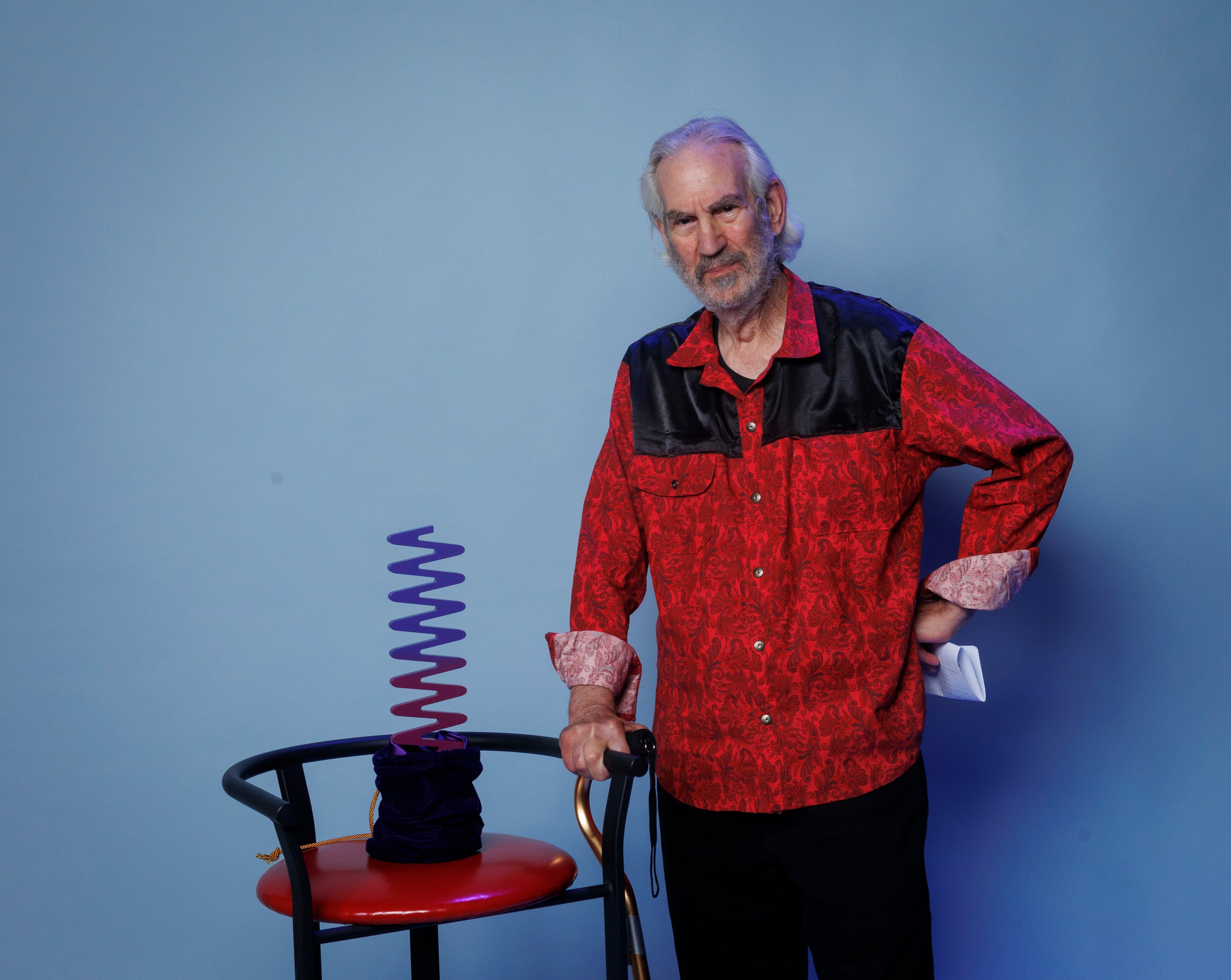 Dave Margoshes - Older white male wearing a red and black, button-down shirt, supporting balance on a walking stick in one hand and the the other hand akimbo. He is standing next to a chair with a blue and purple Ombre wavelength award in a velvet dark blue pouch gathered at the base of the award.