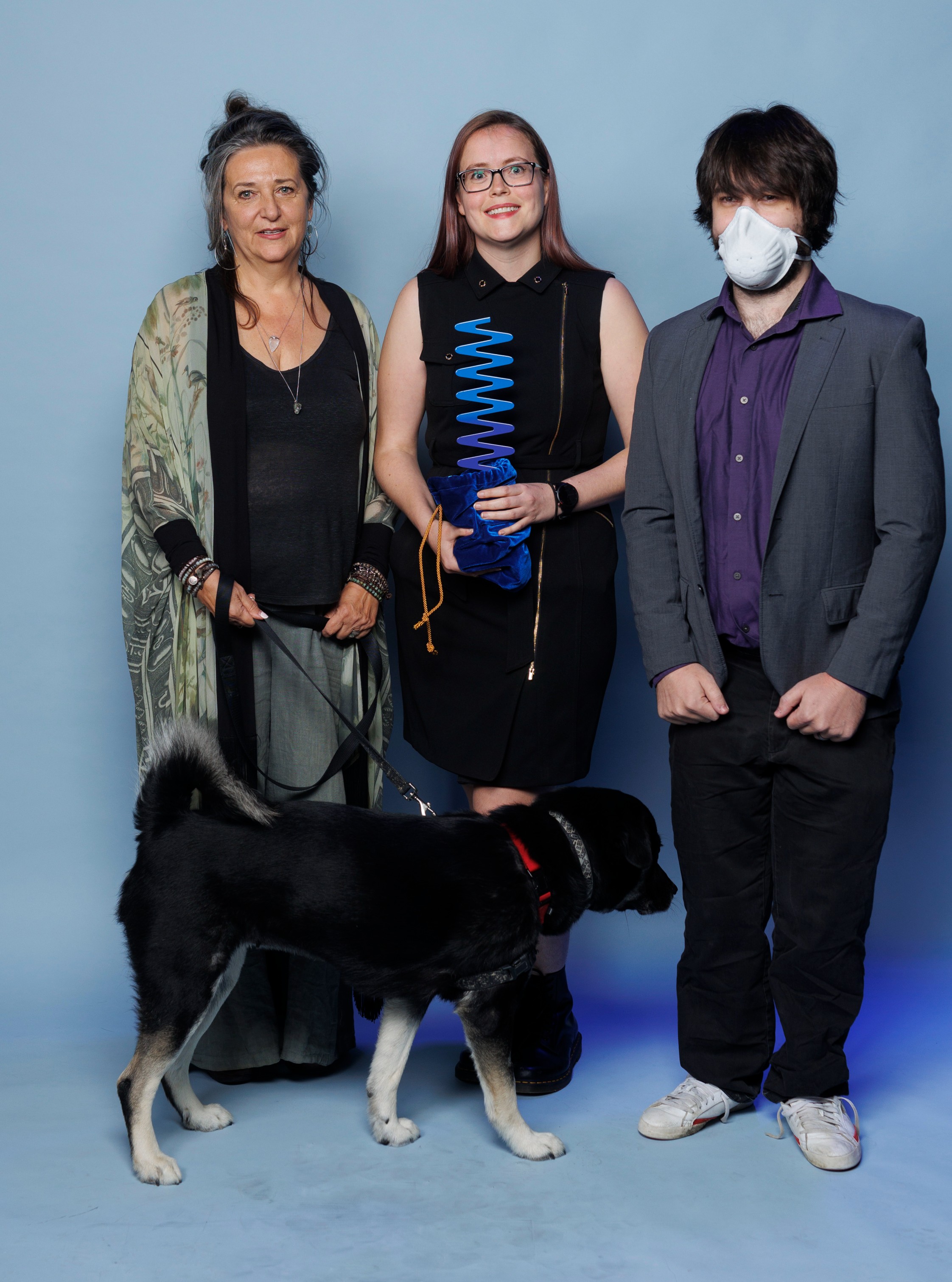 Listen to Dis Team - Photo of three people and a dog. (Left to right description)- The dog is a black, medium-sized service dog with white paws. He is wearing a harness with a red and black collar held by an older woman with gray and black hair pinned up. She is wearing a a balck blouse, grey pants, and grean and grey floral minono. Next to her is a young woman with red hair and balck rimmed glasses in a black dress. She is holding an light blue and blue Ombre wavelength award in a velvet blue pouch gathered at the base of the award. Next to her a young man with medium length hair. He is wearing a mask, a purple shirt, grey jacket, black pants and white sneakers.