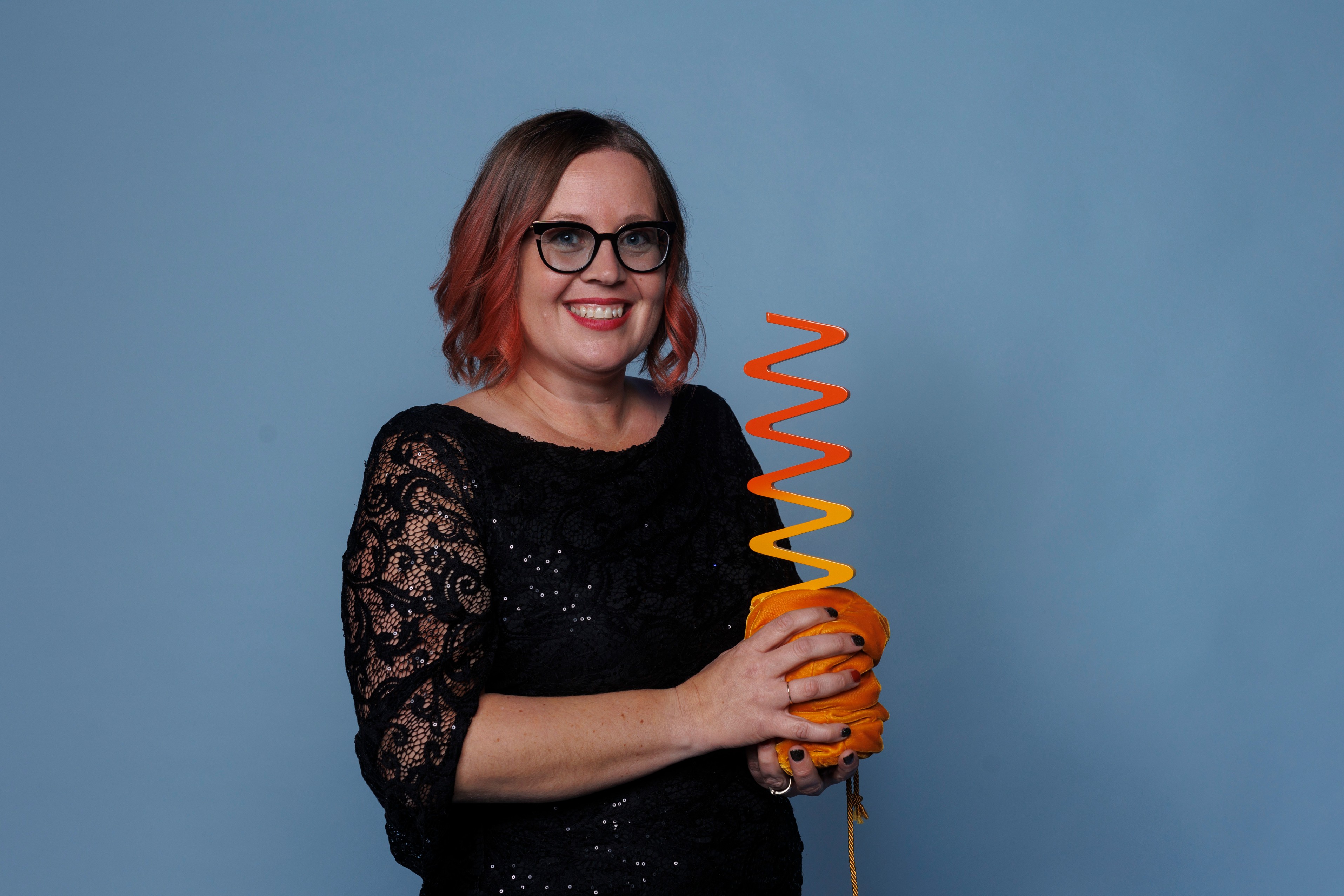 Michelle Grodecki, Arts and Learning recipient - Women black and red Ombre hair, black rimmed and black transparent lens glasses. She is wearing a black dress with lace sleeves and red lipstick smiling brightly while holding an orange wavelength award in a velvet orange pouch gathered at the base of the award.