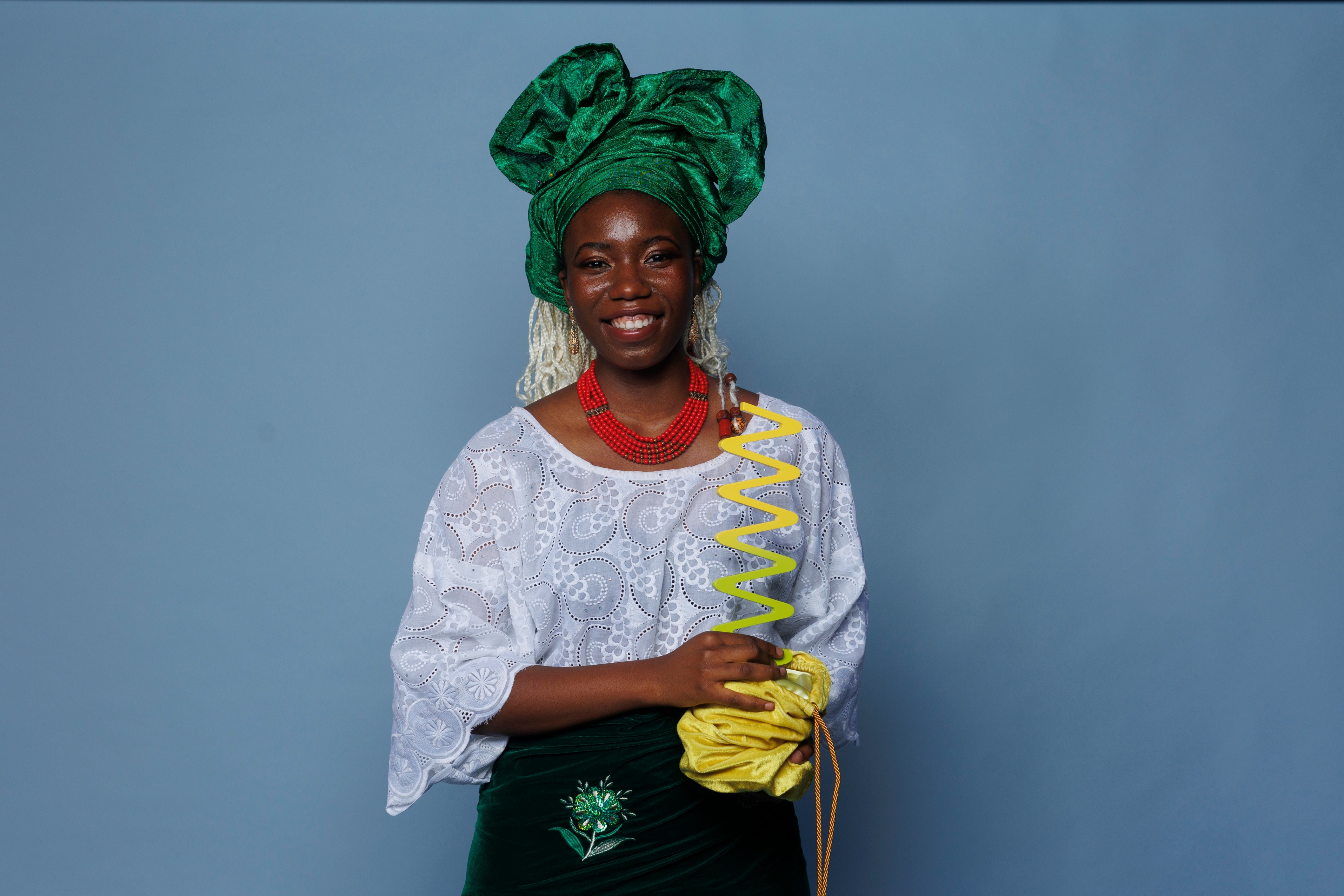 Peace Akintade, young black woman smiling in traditional Nigerian wear - bright green headwrap, large red necklace, white blouse and green skirt. She is holding a yellow wavelength award in a velvet yellow pouch gathered at the base of the award.