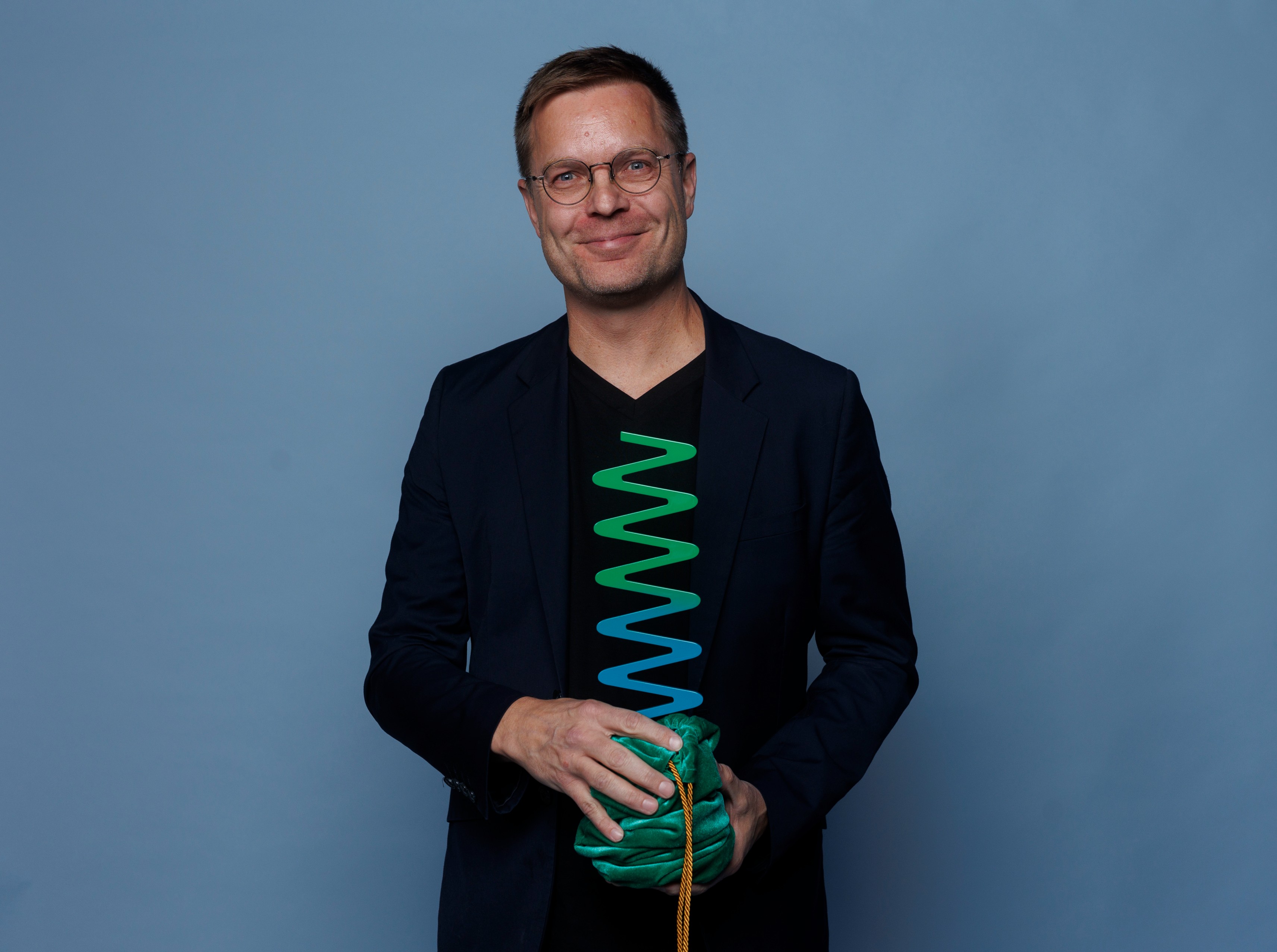 Troy Gronsdahl - White male wearing large, thin-rimmed glasses and a black shirt smiling while holding a green wavelength award in a velvet green pouch gathered at the base of the award.