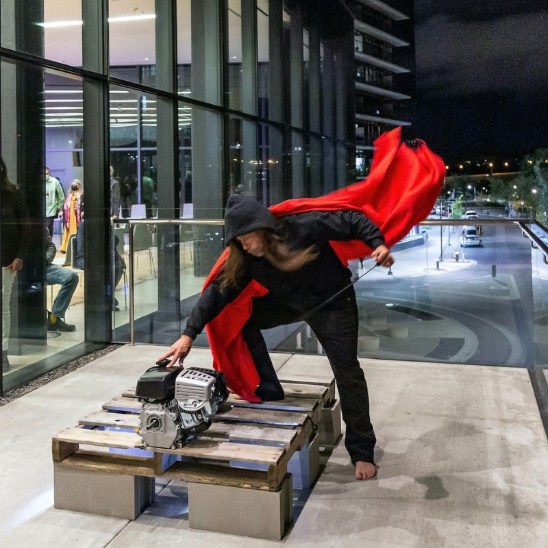 Michel Boutin - Man mid motion wearing black pants and hoodie and red cape. His is barefoot and hunched over pulling a recoil starter rope.