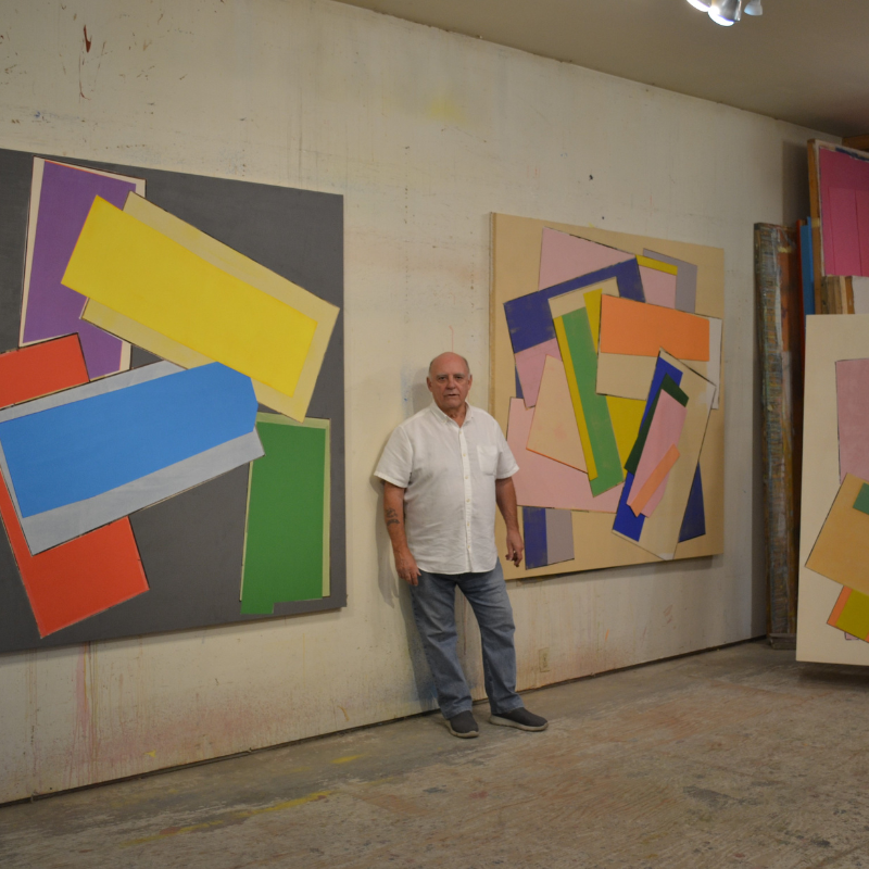 Robert Christie - Man standing in art studio between two large art pieced hung on a wall and other stacked in a corner.
