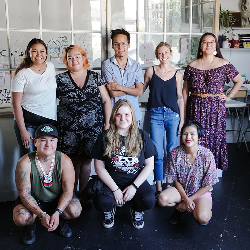 Chokecherry Studios, Saskatoon arts organization. Group of 8 people  in an office space. Description is from left to right at the back - Young indigenous woman with long hair, wearing white t-shirt and jeans smiling broadly with one hand by her side and the other hugging a young woman to her left. Short indigenous young woman with short orange hair wearing a dress. Young indigenous man wearing glasses and a blue button down shirt and his arms folded across his chest with slight smile. White, blonde young woman wearing a black tank top and jeans. Young indigenous woman with long brown hair and wearing glasses smiling. She is wearing a long, off the shoulder floral dress. Description  and then left to right from front - Young indigenous man with tattoos stoops smiling. He is wearing a baseball cap, a dark green tank top and blue jeans shorts. Blonde woman stooped smiling with long blonde hair, black t-shirt with graphics,  jeans and sneakers. She has red makeup around her eyes and is smiling. Young woman in floral top stooping. She is of Asian descent, with short black hair.