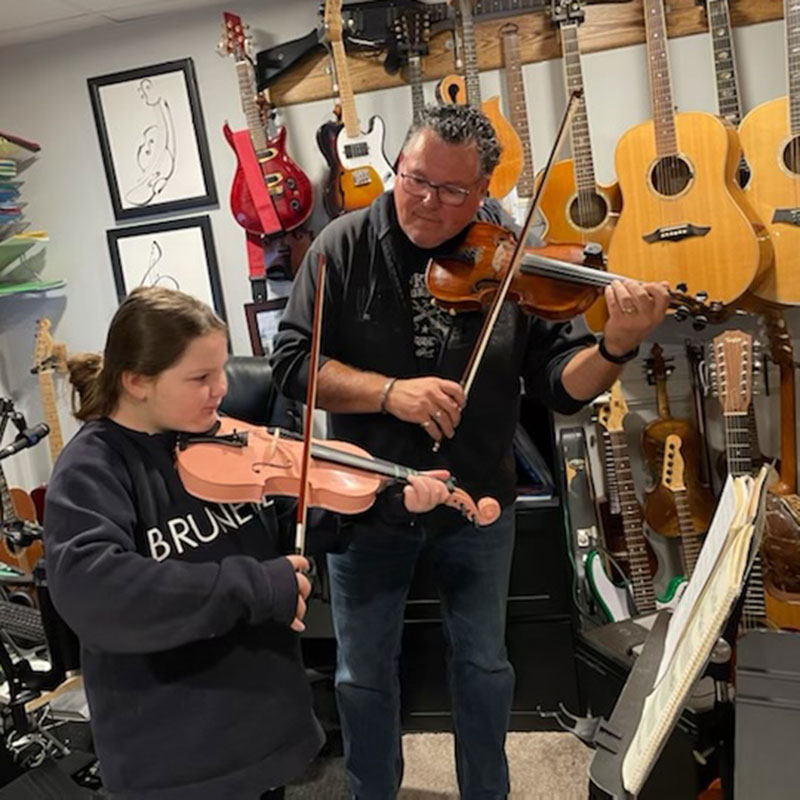 Young child, Hannah Spriggs playing a fiddle in a room filled with instruments. She is being monitored by a man, Donny Parenteau also playing and teaching her to play.