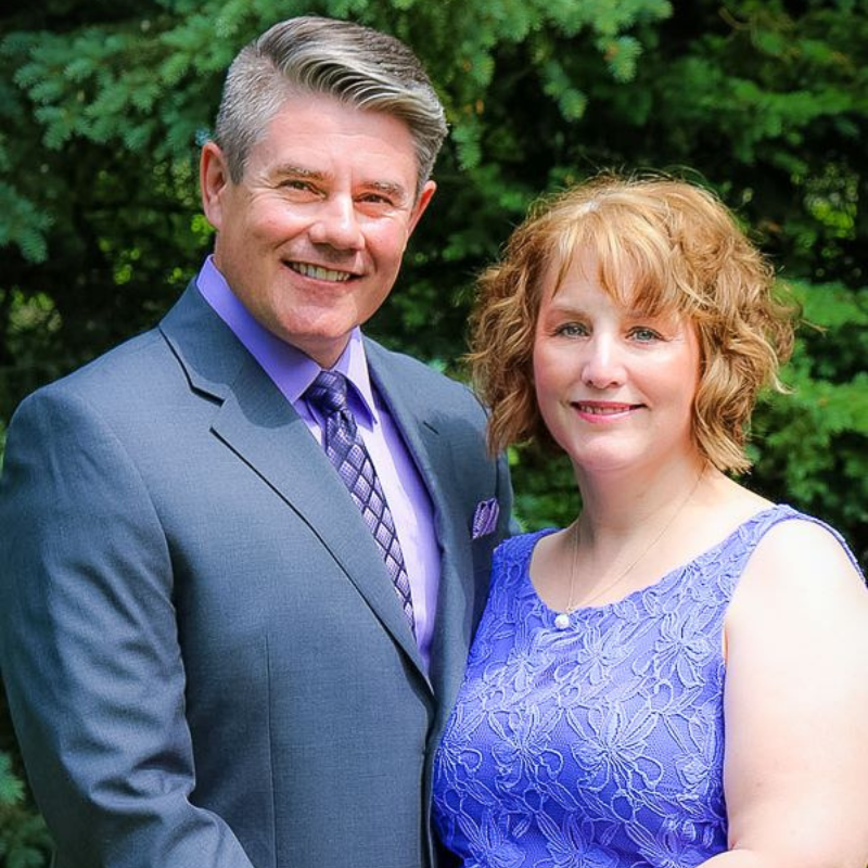 Kevin and Caroll Dyck - Older Man and woman standing closely together. The man has grey hair, is where a grey suit with lavender shirt and purple tie. The woman in wearing a purple dress.