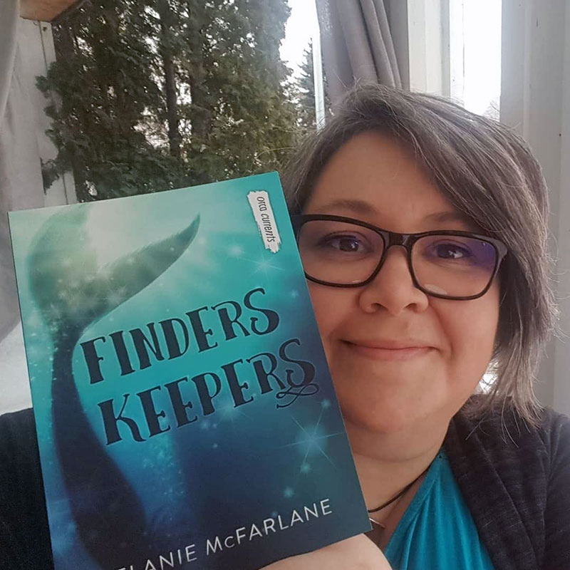 Woman, Melanie McFarlane wearing glasses, smiling and holing a book, titled Finders Keepers next to her face.