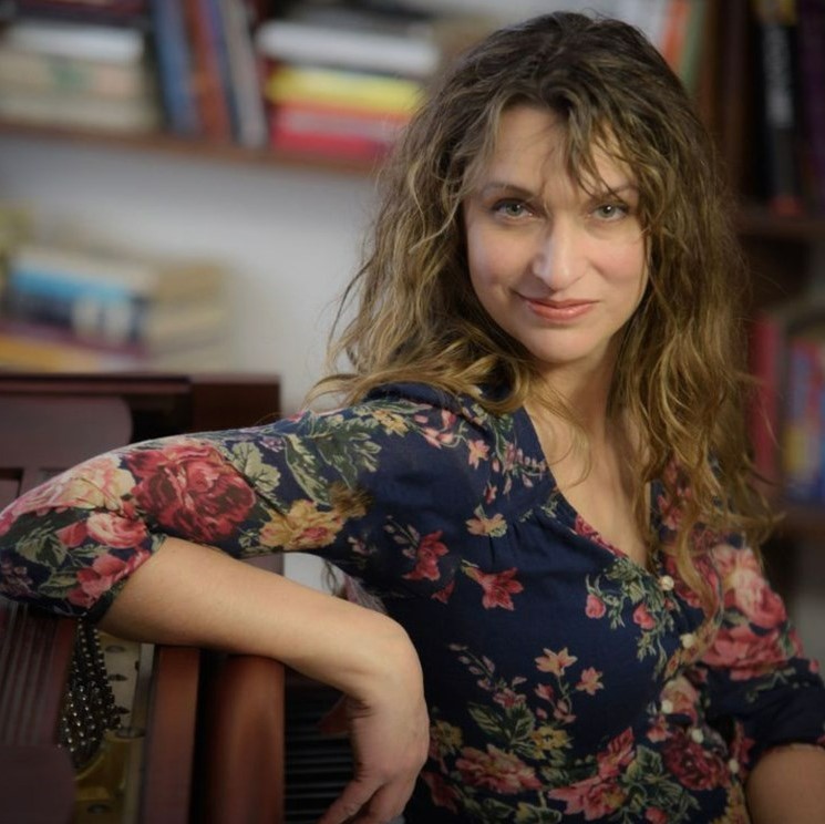 Dara Schindelka - Young woman with brown curly hair with a library behind her.