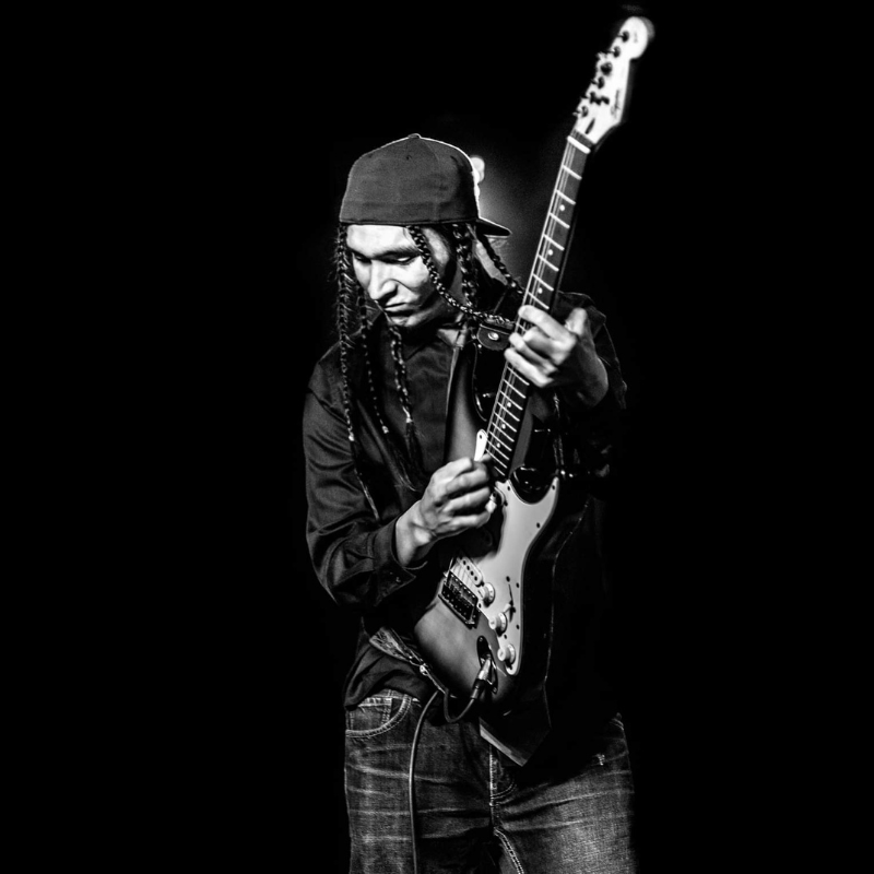 Darion "Dee Cee" Campeau - Indigenous man in black hat, shirt and jeans. He has long plaits in his hair and is holding a guitar. The photo is in black and white. 