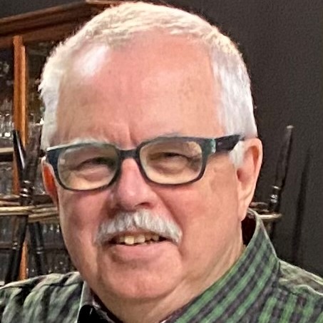 Gordon McCall -  Portrait of older man with gray hair and mustache. He is smiling, wearing glasses and and a green plaid shirt.