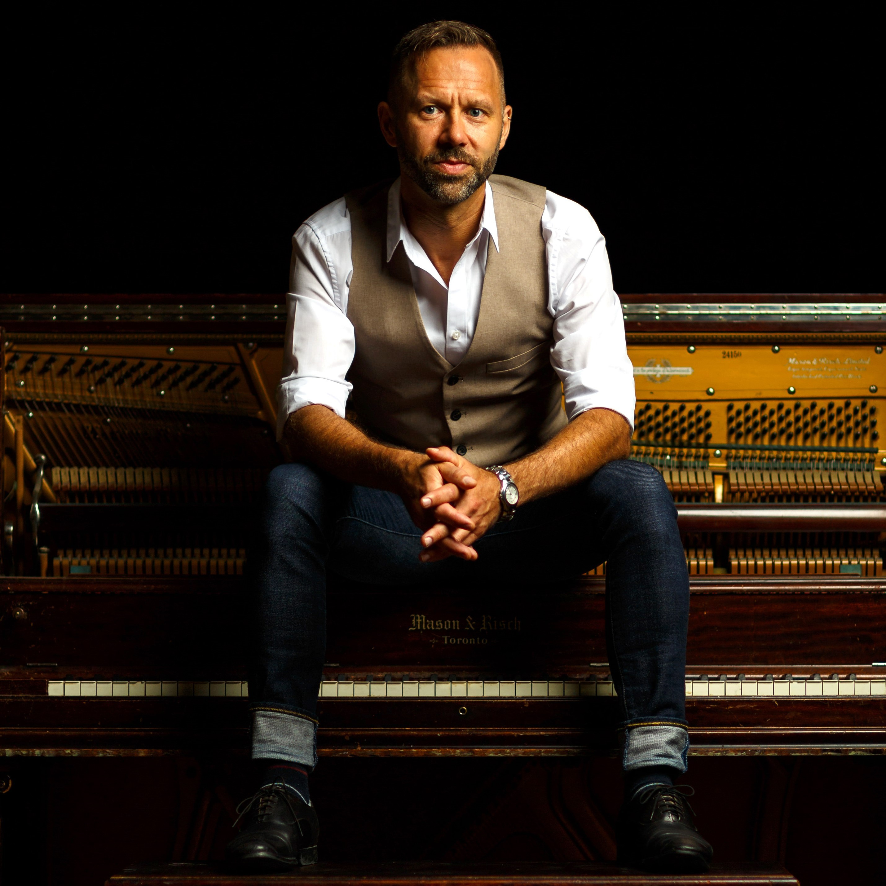 Jeffery Straker - man sitting on a vintage piano wearing a white shirt, brown vest, blue jeans and black boots.