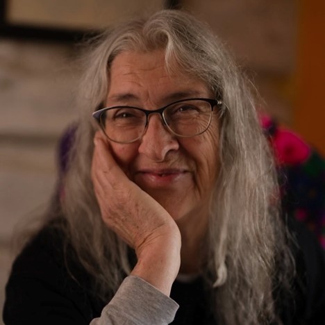 Marjorie Beaucage, SK Arts 75th anniversary nominator - Métis woman with long grey hair wearing glasses and smiling with one hand on her cheek.