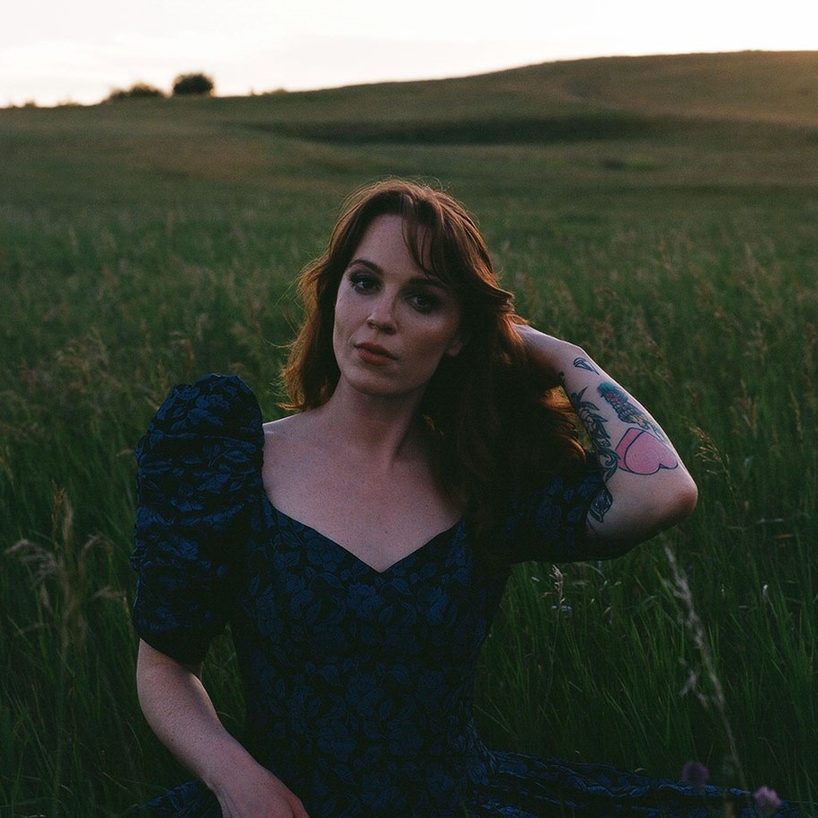 Megan Nash - SK Arts 75th anniversary nominator - Photo by Gina Brass, shows a women in a field with red hair, She is wearing a blue dress with big sleeves and had one arm covered in tattoos going through her hair.