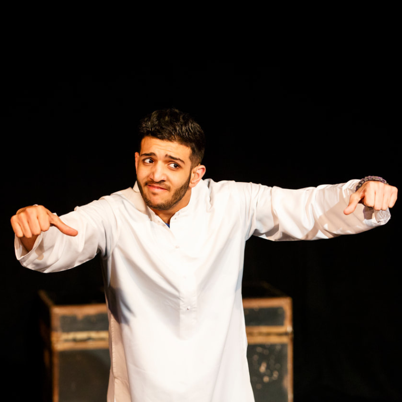 Mustafa Alabssi - Young man wearing to a long with shirt with longs sleeves on stage mid-performance with arms outstretched.