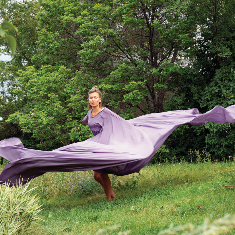 Robin Poitras - Older woman mid-dance on a hill with trees behind her. Her long lavender dress is moving with her in the wind.