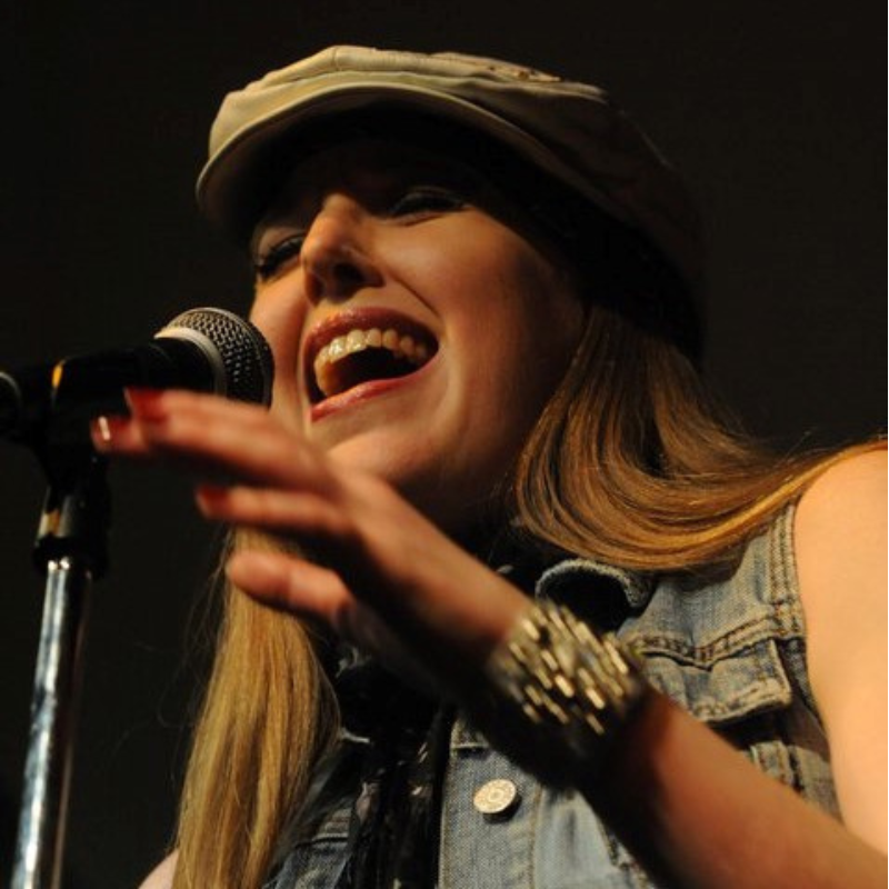 Tara Dawn Solheim - Woman wearing a hat and sleeveless jean jacket singing into a microphone