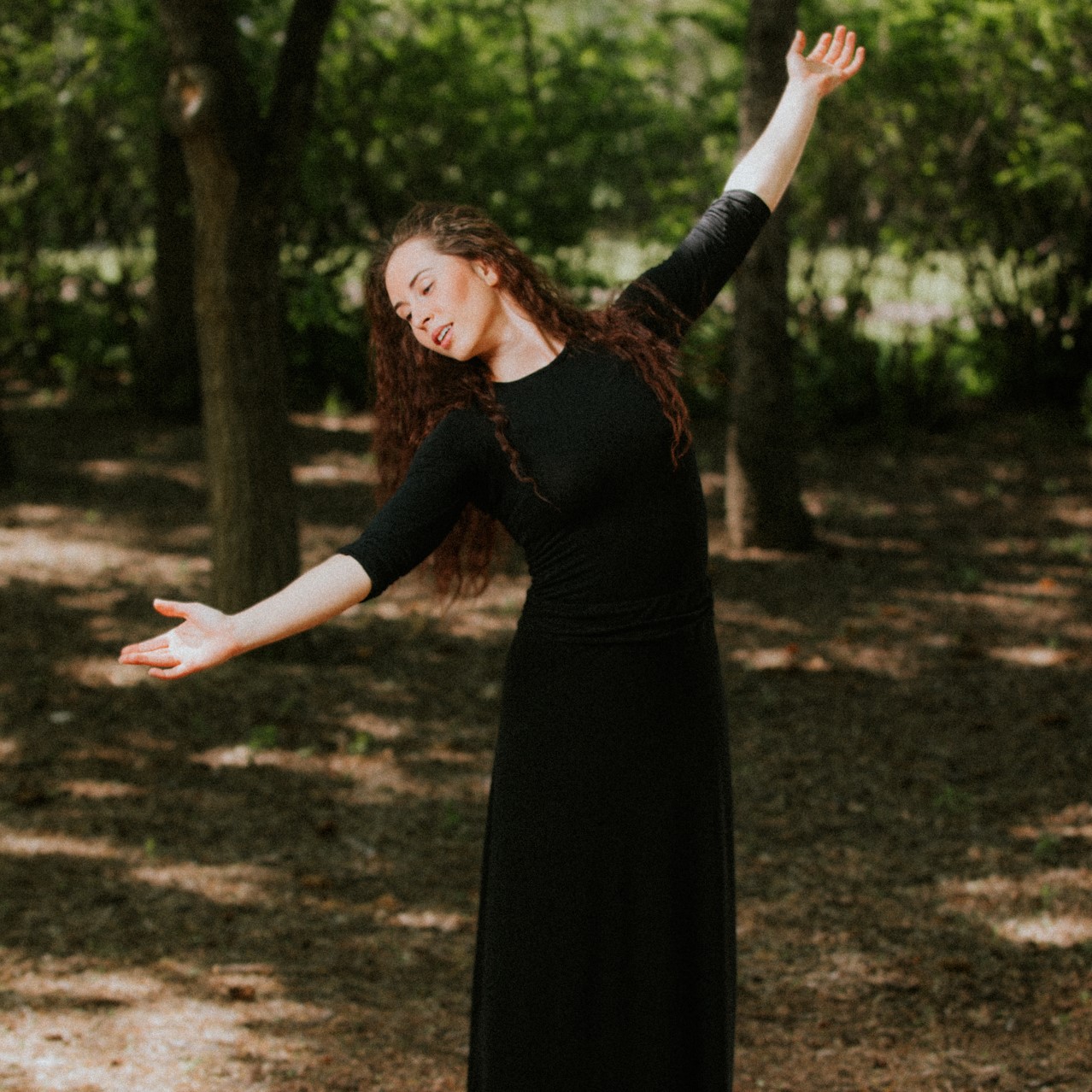 Tessa Rae Kuz - Young woman in long black dress in the woods mid-dance.