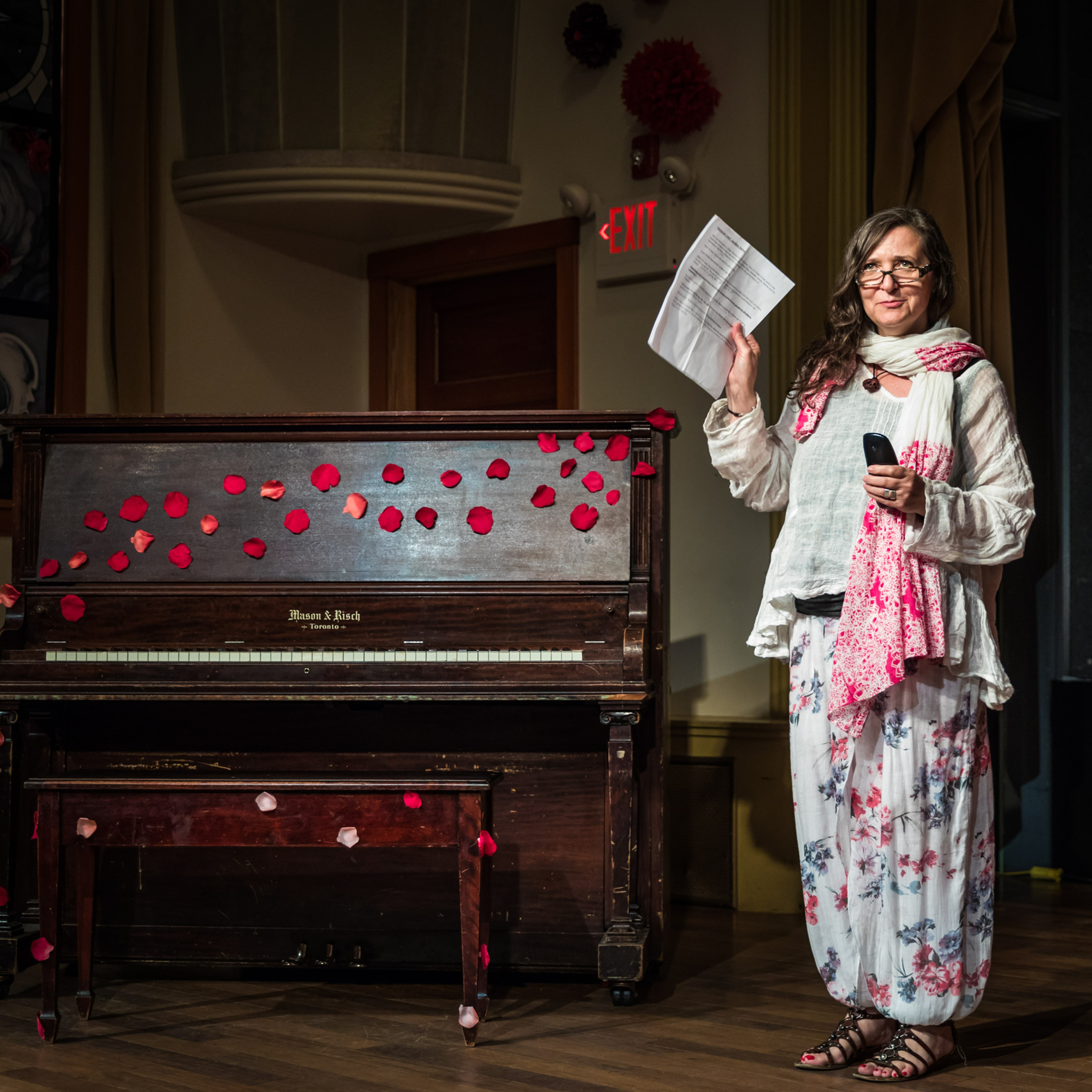 Traci Foster - Woman wearing glasses  on stage wearing a long sleeve top, a long skirt and a  scarf. She is holding up a sheet of paper and standing next to a piano.
