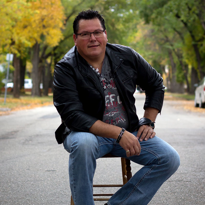 Indigenous man sitting on a stool in the middle of a street lined with homes and trees. He is wearing a leather jacket, a grey t-shirt with graphics and blue jeans. He has one hand on his leg and the elbow of the other hand on his thigh.