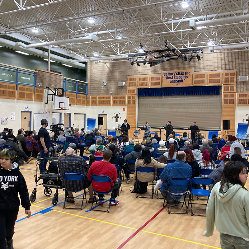 Image of gathering in a basketball court. There are chairs facing a stage with 4 speakers.