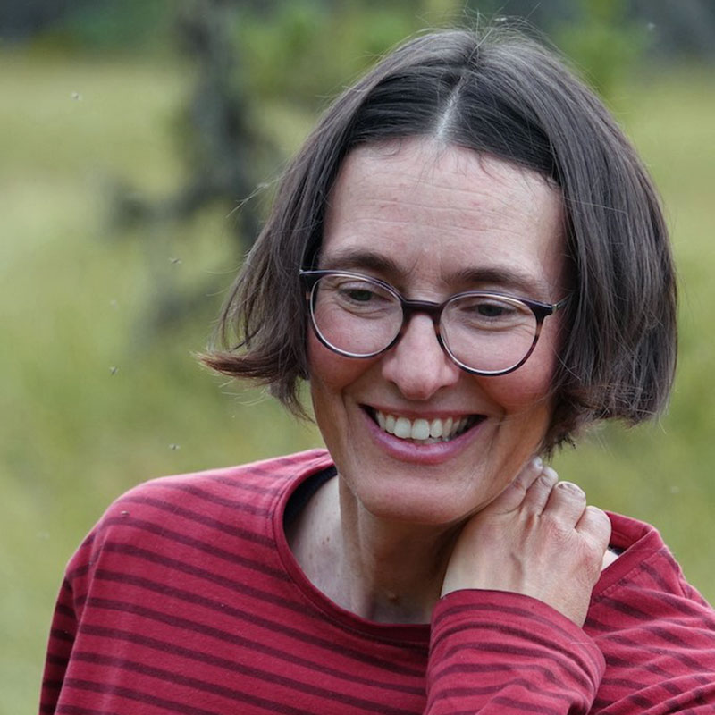 Miriam Körner - Indigenous woman with a short brown bob haircut. She is wearing glasses, a red and black striped long sleeve shirt and smiling vroadly. The background is greenery that is blurred.