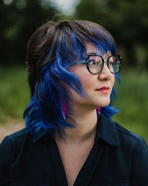 Portrait of white presenting woman with blue hair and wearing a black short and black rimmed glasses. Courtney Bates-Hardy 
