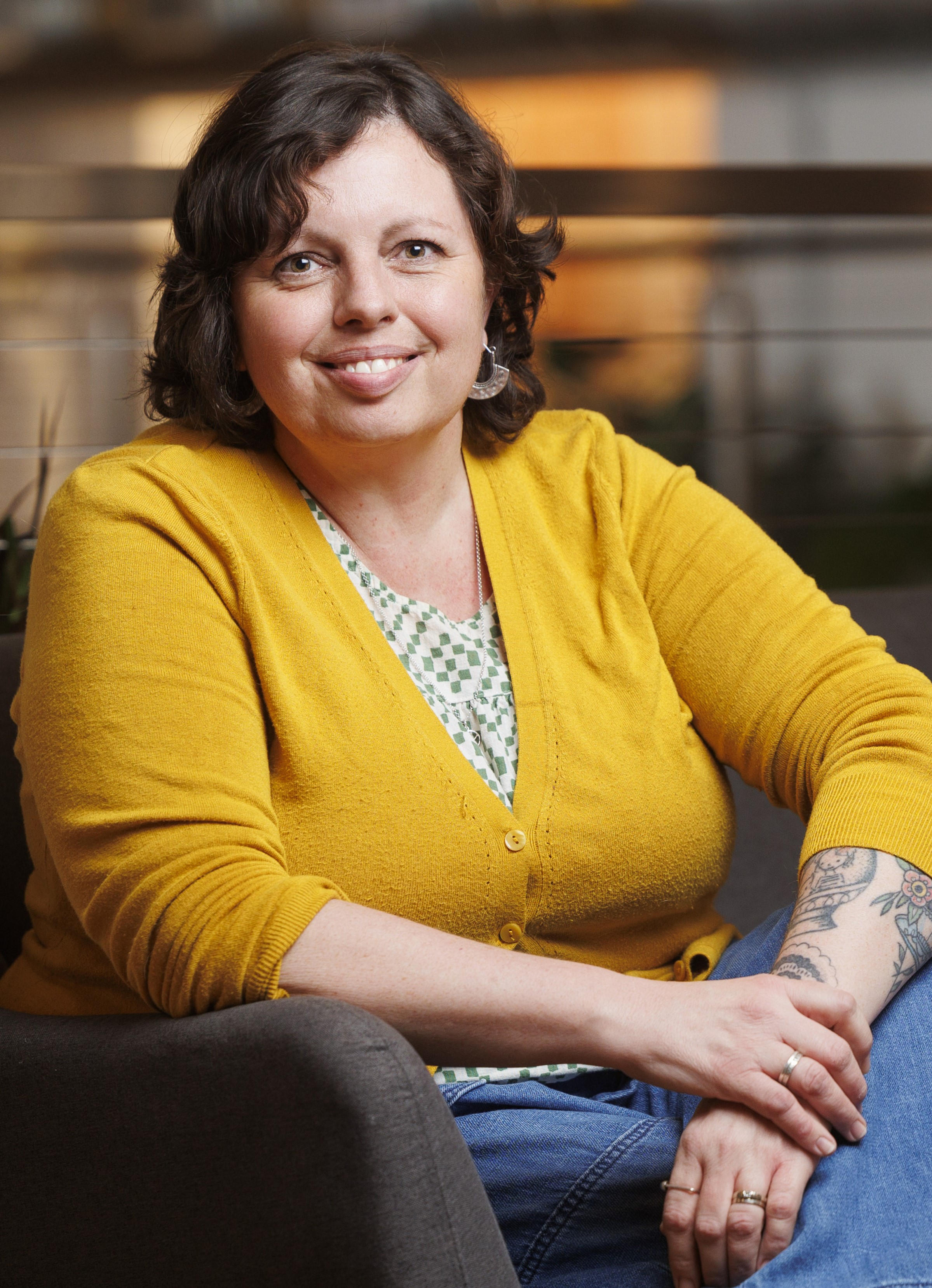 Carole Epp- SK Arts Board Member, Canadian artist, Ceramicist - A woman with short brown hair wearing a mustard colour cardigan over a floral blouse smiling with on leg across her knee.