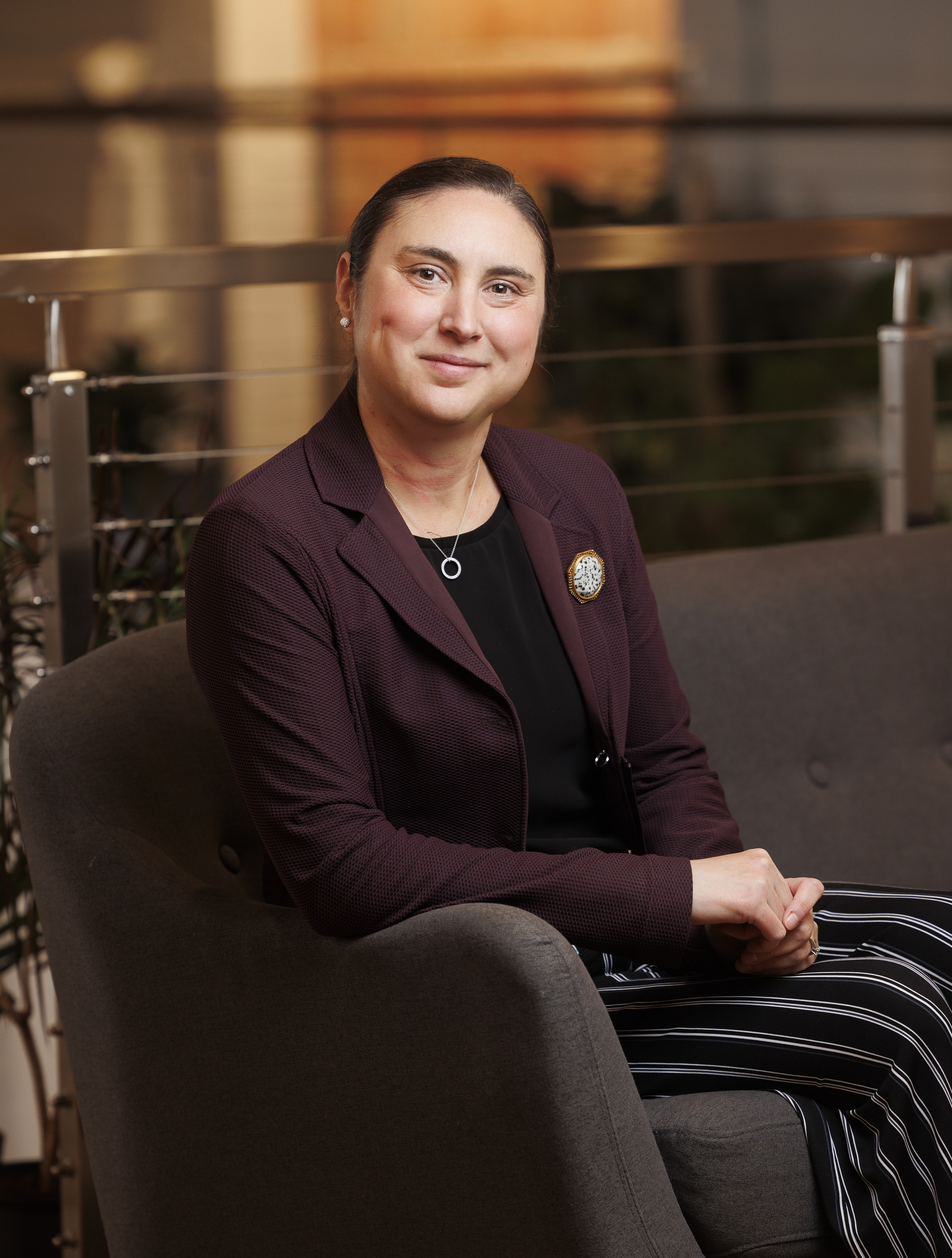 Diana Domm Smythe - SK Arts Board Member, Management professional - Portrait of a woman with her hair back, and deep dimples smiling. She is wearing a burgundy blazer jacket with a black blouse and wearing a broach, and striped slacks sitting in an arm chair.