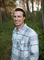 Keith Busch – SK Arts Board Member - Tall, white male with brown hair wearing a blue shirt, black vest and grey pants. He is smiling, is in a forest environment, and wearing a blue/grey lard plaid print shirt.