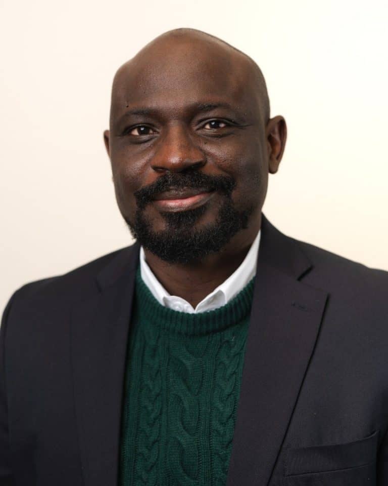 Michael Afenfia - SK Arts board member - Portrait of bald, black man with a mustache smiling. He is wearing a  white shirt with a collar, a dark green sweater and black jacket.