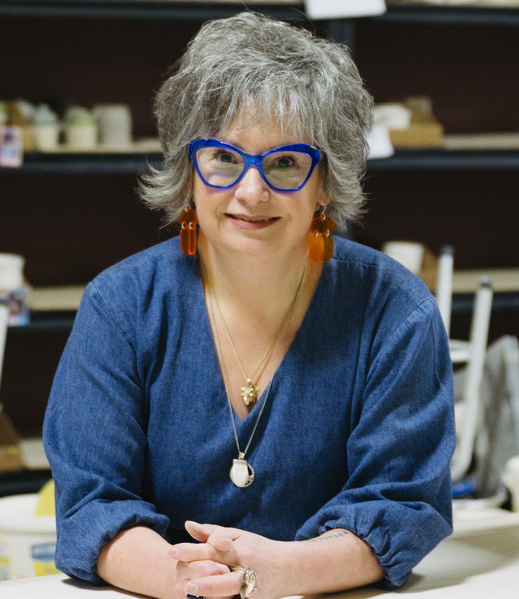 Cheryl Ring- Visual Artist - Portrait of woman with short grey hair and blue rimmed glasses, orange hanging earrings and blue blouse with ling sleeves leaning over and smiling,