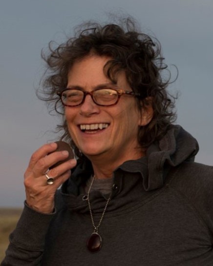 SK Arts board member and Saskatchewan artist Barbara Meneley. Headshot of woman with short curly hair blowing in the wind. She is wearing coloured sunglasses and smiling broadly.