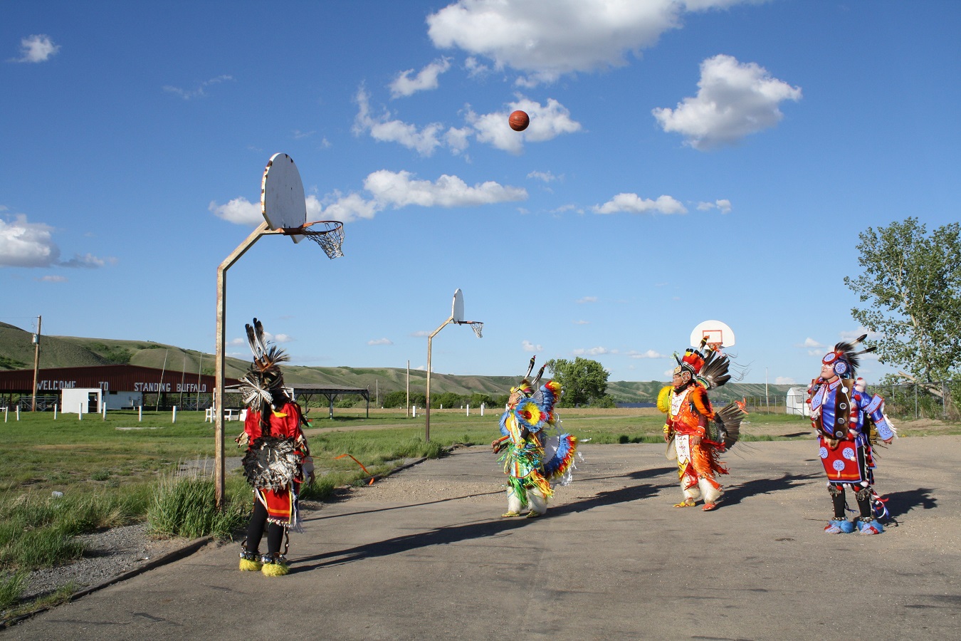 Four Indigenous men dressed in traditional regalia play basketball outside.