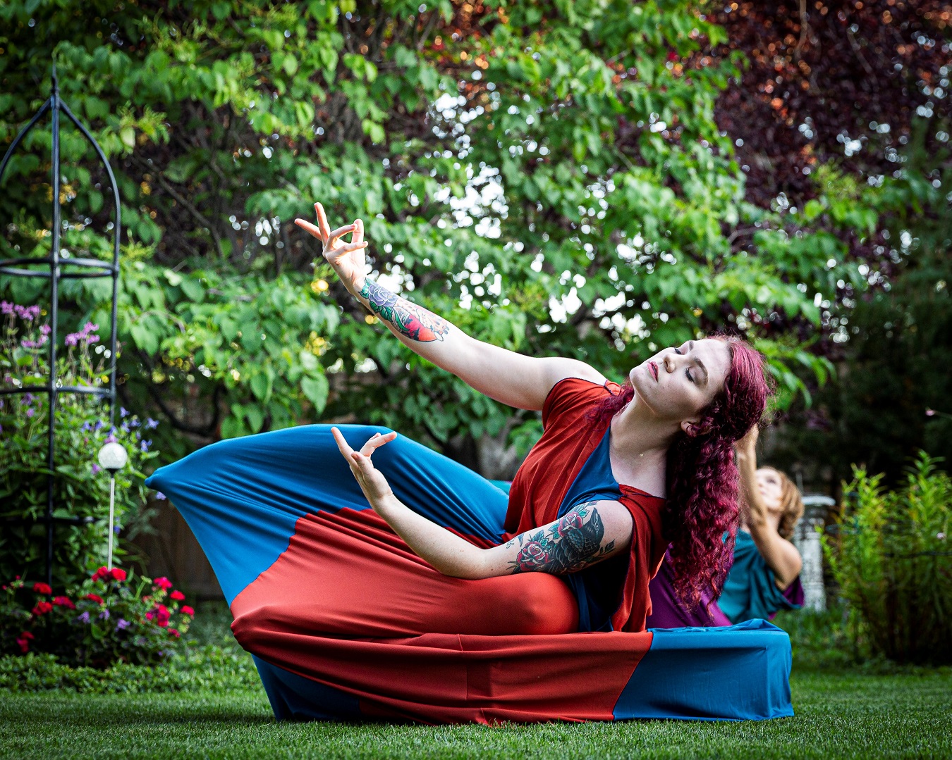 A woman wearing red and blue extends both of her arms while sitting on a trampoline.