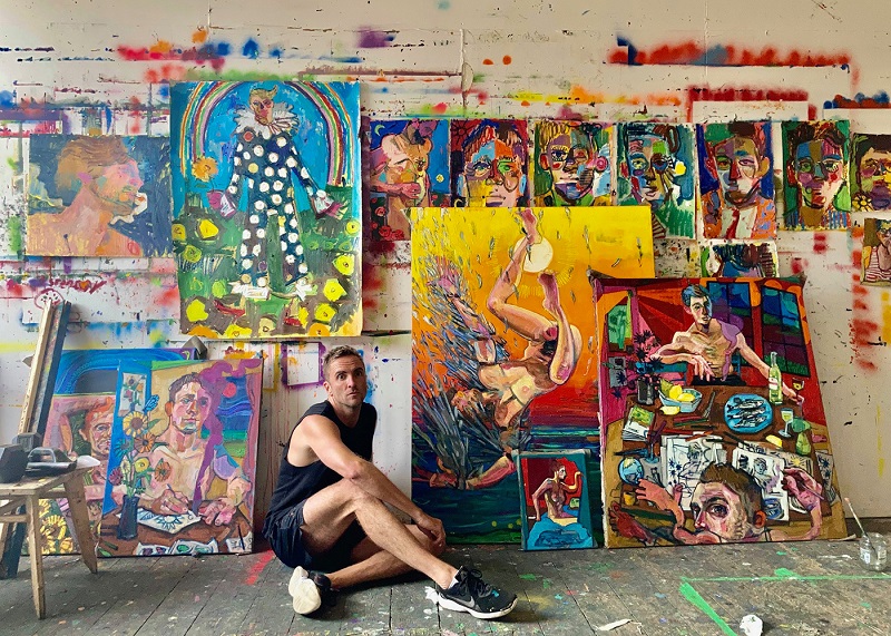 Andrew Salgado sitting in his studio in front of several of his colourful paintings