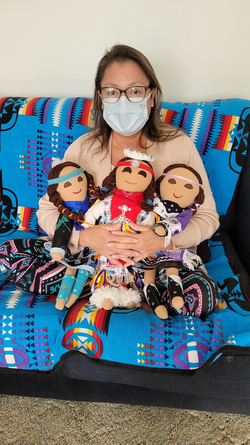 Elaine McArthur sitting on a sofa, wearing a mask, and holding three dolls that are dressed in traditional regalia