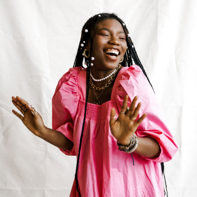 eace Akintade, black young woman in a pink dress and braids, with a big smile touching her forehead and holding up both hands in candid pose