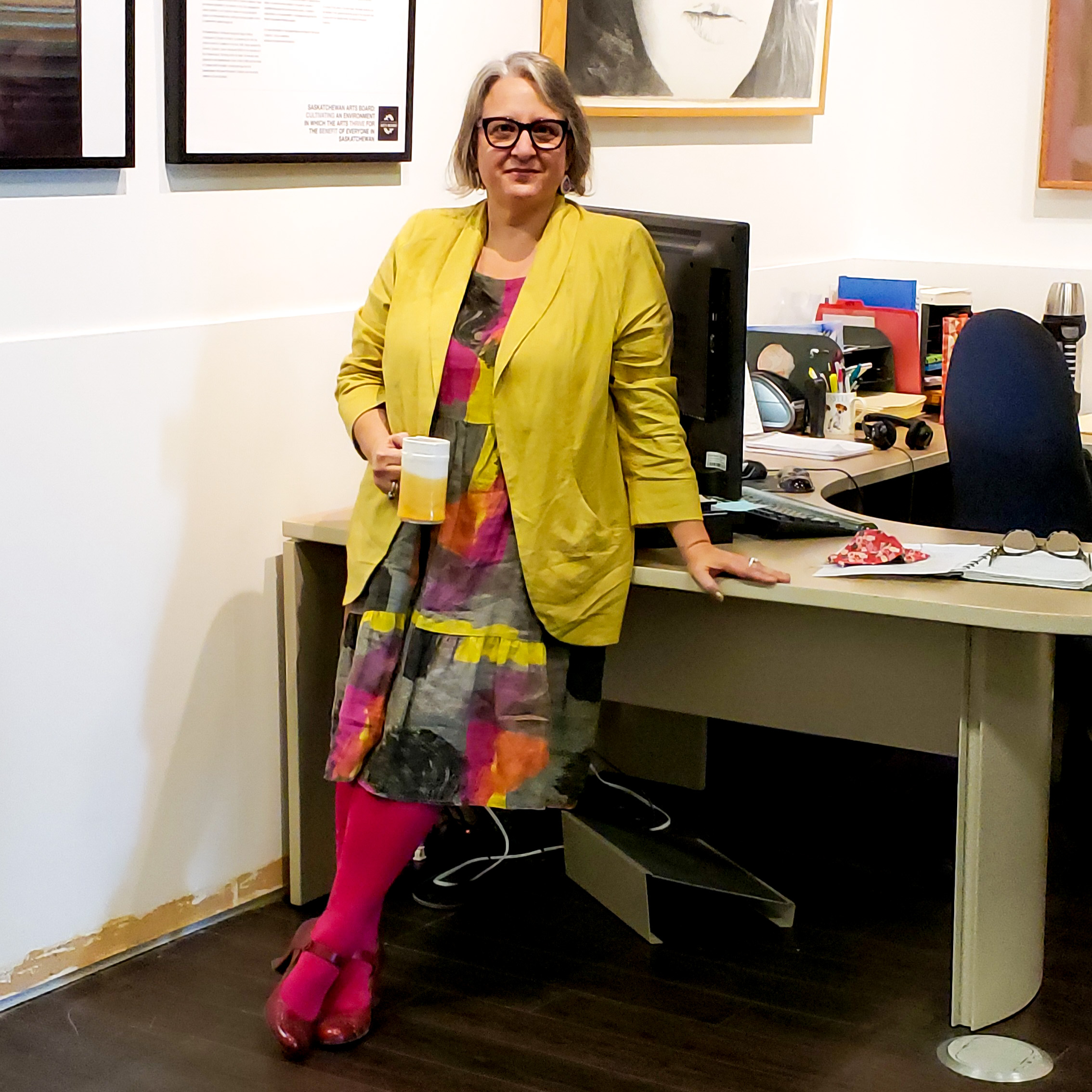 Belinda Harrow, SK Arts Permanent Collection Consultant - Older woman leaning on a desk with crossed legs holding a coffee mug. She has grey hair, wearing glasses and smiling. She is wearing a mustard yellow jacket, a colourful dress, red stockings and strapped shoes.