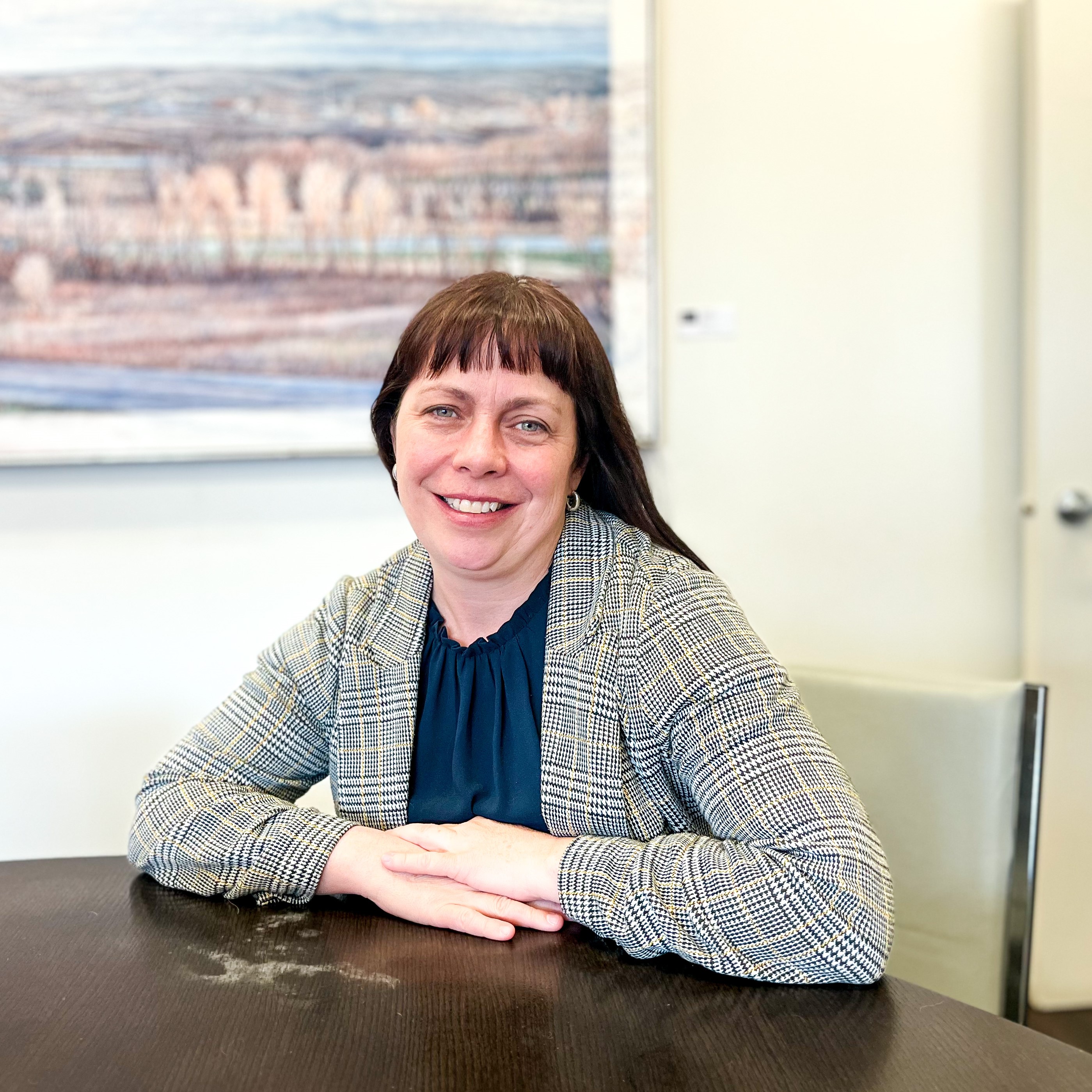 Jody Greenman-Barber, Program Consultant - White woman in an office. She has brown hair, bangs, is wearing a plaid jacket and black blouse. There is a painting behind her,