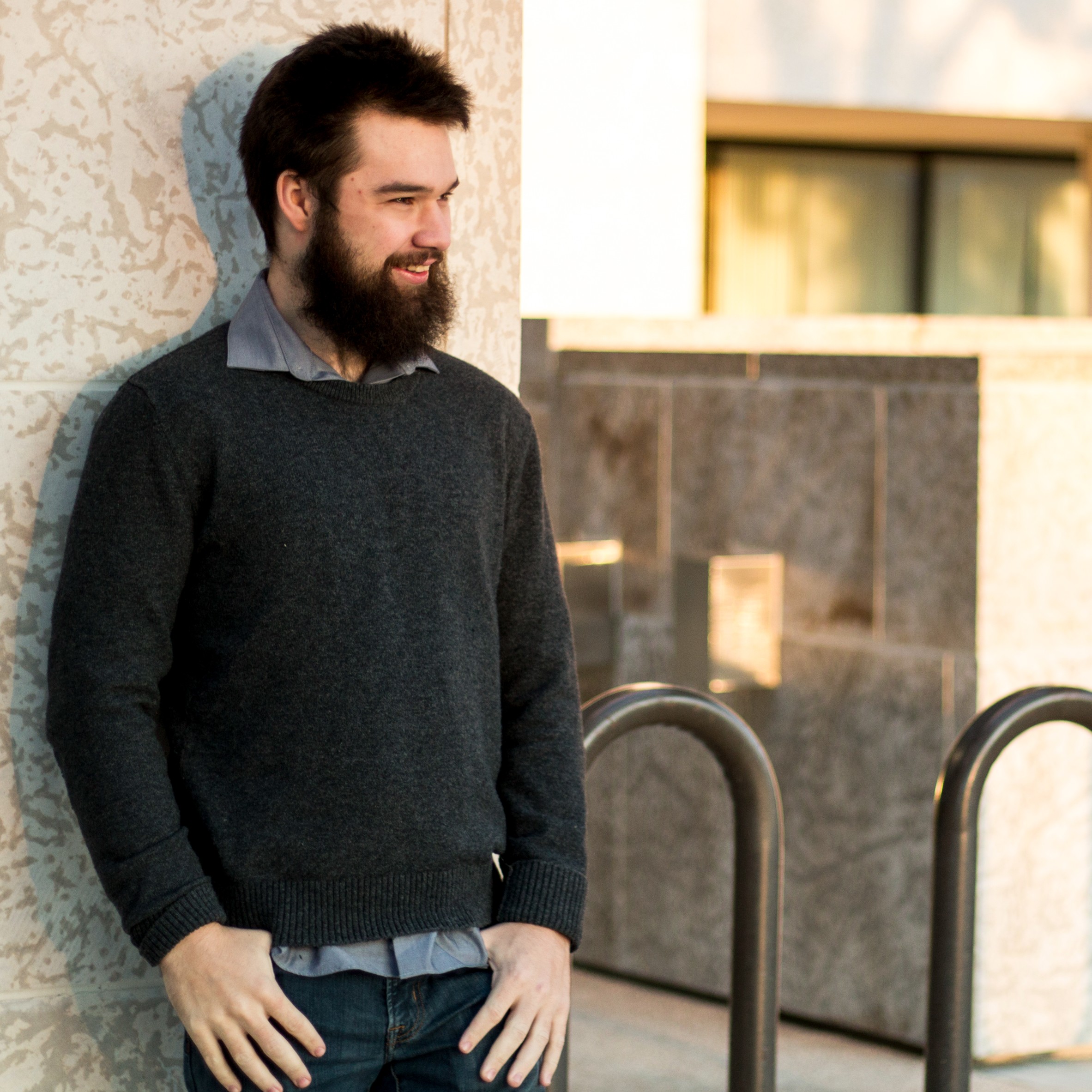 John Loeppky- White make leaning against a wall. He as short hair, a beard and wearing a plaid collar shirt with a black sweater over it and jeans, His thumbs are hooked in his pockets.