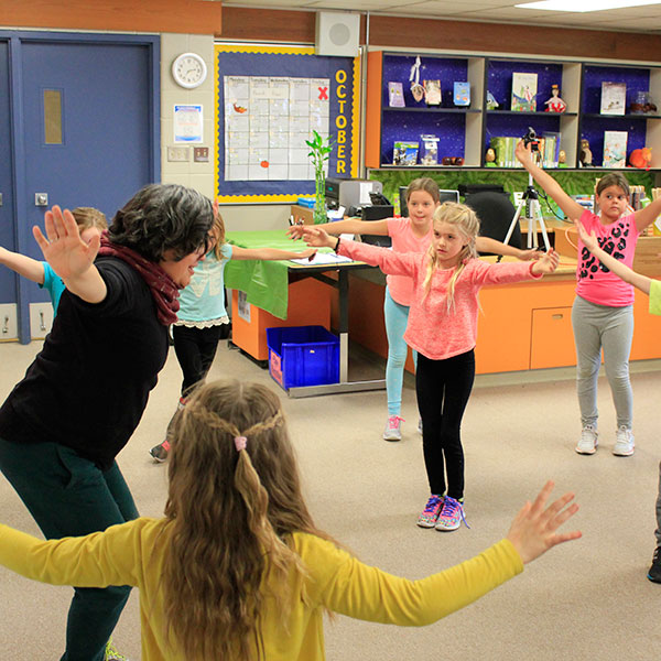 Artist standing in a circle in a classroom with 9 students. They all have their knees together and bent, with their arms stretched wide. The teacher looks on from the back of the classroom.