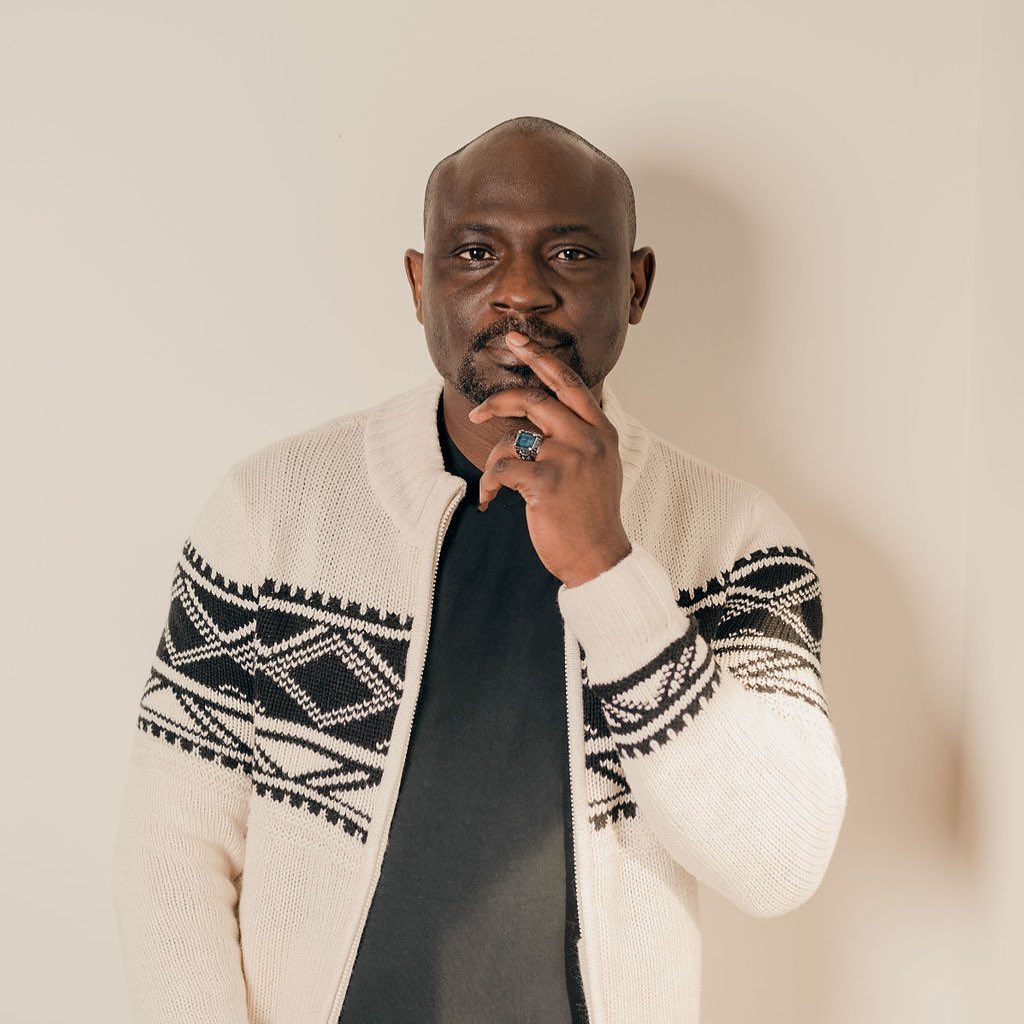 Michael Afenfia, Nigerian Canadian author. Portrait with cream coloured background of black Black man wearing a cream and black sweater and a black shirt. He has one finger loosely on his lips.