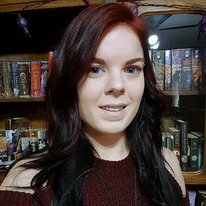 Vanessa Wraithe, Administrative Coordinator - White woman with red and black hair. She is smiling and is in front of a bookcase.