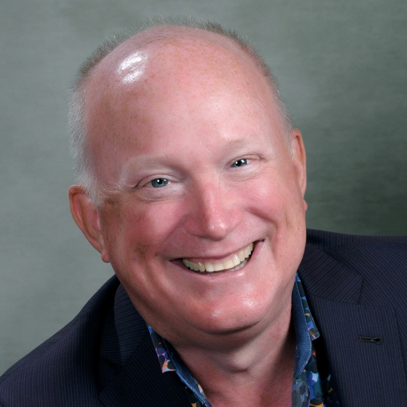 Headshot of Michael Jones, CEO of SK Arts. Older while male hearing navy blue jacket and navy blue patterned short smiling broadly.