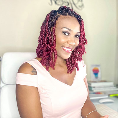 Shandean Reid, Communications Strategist - Black woman with black and red braids. She is wearing a light pink dress and smiling.