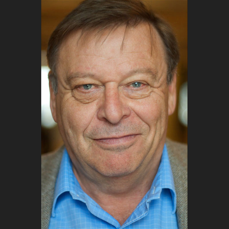 Guy Vanderhaeghe - Close up profile of older man. He is smiling slightly, wearing a baby blue shirt and grey jacket.
